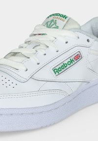 Fehér Reebok Classic bőr sportcipő zöld logóval és Union Jack zászlóval az oldalon, fehér fűzőkkel és texturált fehér talppal.