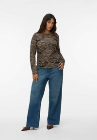 Haut à manches longues avec un motif de rayures brunes et noires en ondulation, associé à un jean en denim bleu à jambes larges. Chaussures brunes à bout pointu avec des accents.
