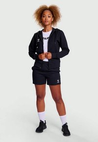 Svart hettejakke med glidelås og logo, sammen med en hvit t-skjorte og svarte shorts. Modellen har på seg svarte sko og hvite ankelsokker.