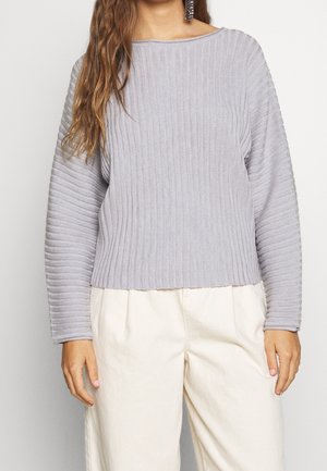 Pull en maille grise avec un large col et des manches longues, présentant un tissu texturé. Assorti à un pantalon clair et ample.