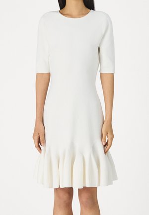 Woman wearing a white knee-length dress with elbow-length sleeves and a flared, pleated hem.