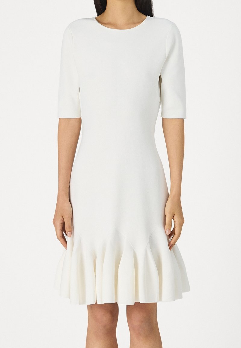 Femme portant une robe blanche arrivant aux genoux avec des manches jusqu'aux coudes et un ourlet évasé et plissé.