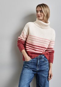 Maglione lavorato a maglia con un colletto alto a coste in crema e rosso, caratterizzato da strisce orizzontali in tonalità di rosa. Abbinato a jeans in denim blu.