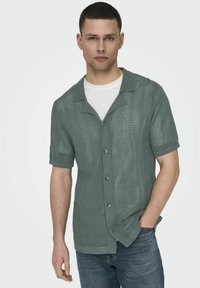 Kurzarmiges, grünes Strickhemd mit Kragen, aus strukturiertem Stoff, mit fünf Knöpfen und umgeschlagenen Ärmeln, über einem weißen T-Shirt getragen.