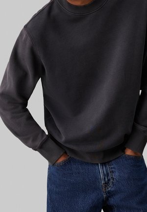 Sudadera gris oscuro con cuello redondo y puños acanalados, llevada sobre unos jeans de mezclilla azules, que muestran un corte relajado y una textura suave.