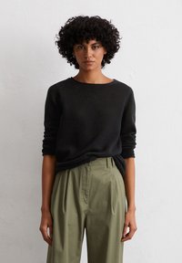 Schwarzer, strukturierter Pullover mit rundem Ausschnitt, kombiniert mit olivgrünen, gefältelten Hosen. Die Ärmel des Pullovers sind hochgekrempelt. Minimale Accessoires.