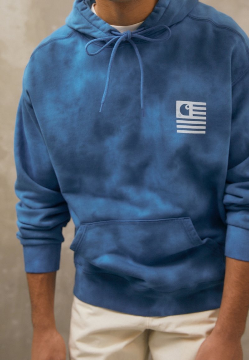 Personne portant un sweat à capuche tie-dye bleu avec une poche avant et un motif blanc sur la poitrine, associé à un pantalon clair.