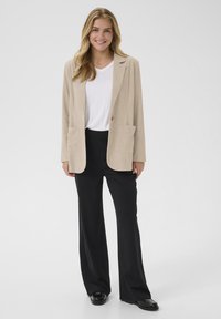 Beige corduroy-takki kahdella etutaskulla ja yhdessä napilla, yhdistettynä mustiin leveisiin housuihin. Asusteena valkoinen t-paita.