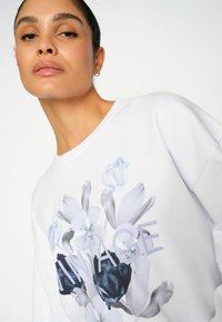 Sudadera blanca con un estampado floral en tonos de azul y negro, con la palabra "VINTAGE" integrada en el diseño.