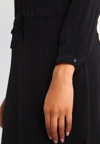 Czarna sukienka z długimi rękawami, wykonana z lekkiego materiału. Posiada detal w postaci wiązania w talii oraz mankiety zapinane na guziki. Miękka tekstura, prosta sylwetka.