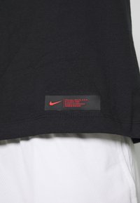 Czarna bawełniana koszulka z czerwonym metkami Nike. Metka zawiera informacje o założeniu i szczegóły konferencji. Koszulka ma gładką teksturę.