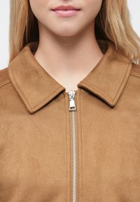 Tan faux ruskindjakke med spids krave, sølv lynlås og glat tekstur, med fokus på den øvre torso.