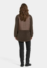 Bruine fleecejack met een gewatteerde, bruine panel op de achterkant. Het heeft lange mouwen en een afgeronde zoom, gecombineerd met zwarte slim-fit jeans en laarzen.
