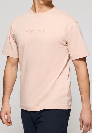 Mand iført en lyserød T-shirt med korte ærmer med præget tekst "Polo Club" og mørke bukser mod en ensfarvet baggrund.