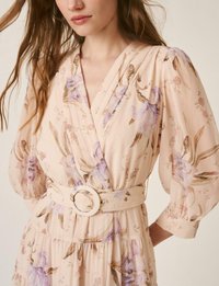 Mujer con un vestido cruzado floral beige con mangas abullonadas y un cinturón ancho con una hebilla redonda grande.