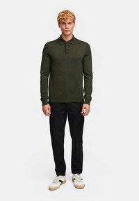 Donkergroen poloshirt met lange mouwen en een geribbelde textuur, voorzien van een kraag met drie knopen, gecombineerd met zwarte slim-fit broek en witte sneakers.