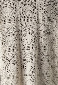 Pletená tkanina v krémové barvě s komplikovaným krajkovým vzorem zahrnujícím hexagonální tvary a motivy diamantů, ukazující jemné, texturované detaily po celém povrchu.