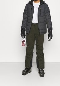 Svart puffjacka med gula detaljer, grå skjorta, olivgröna byxor, svarta handskar, och skidhjälm med rosa linser hålls i handen.