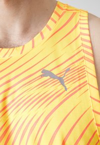 Žlutý sportovní tílko s červenými a oranžovými diagonálními pruhy. Obsahuje šedé logo Puma na hrudi a texturovaný vzor látky.