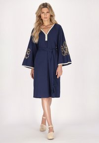 Rochie de culoare albastră închisă cu mâneci clopot, având broderie aurie pe mâneci, un decolteu în V și o centură cu ciucuri. Textură netedă și ușoară.