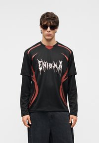 Νεαρός άνδρας που φορά μαύρο μακρυμάνικο πουκάμισο με κόκκινες λεπτομέρειες και λευκή επιγραφή «ENIGMA», σκούρα γυαλιά ηλίου και σκούρο παντελόνι, στέκεται μπροστά σε λευκό φόντο.