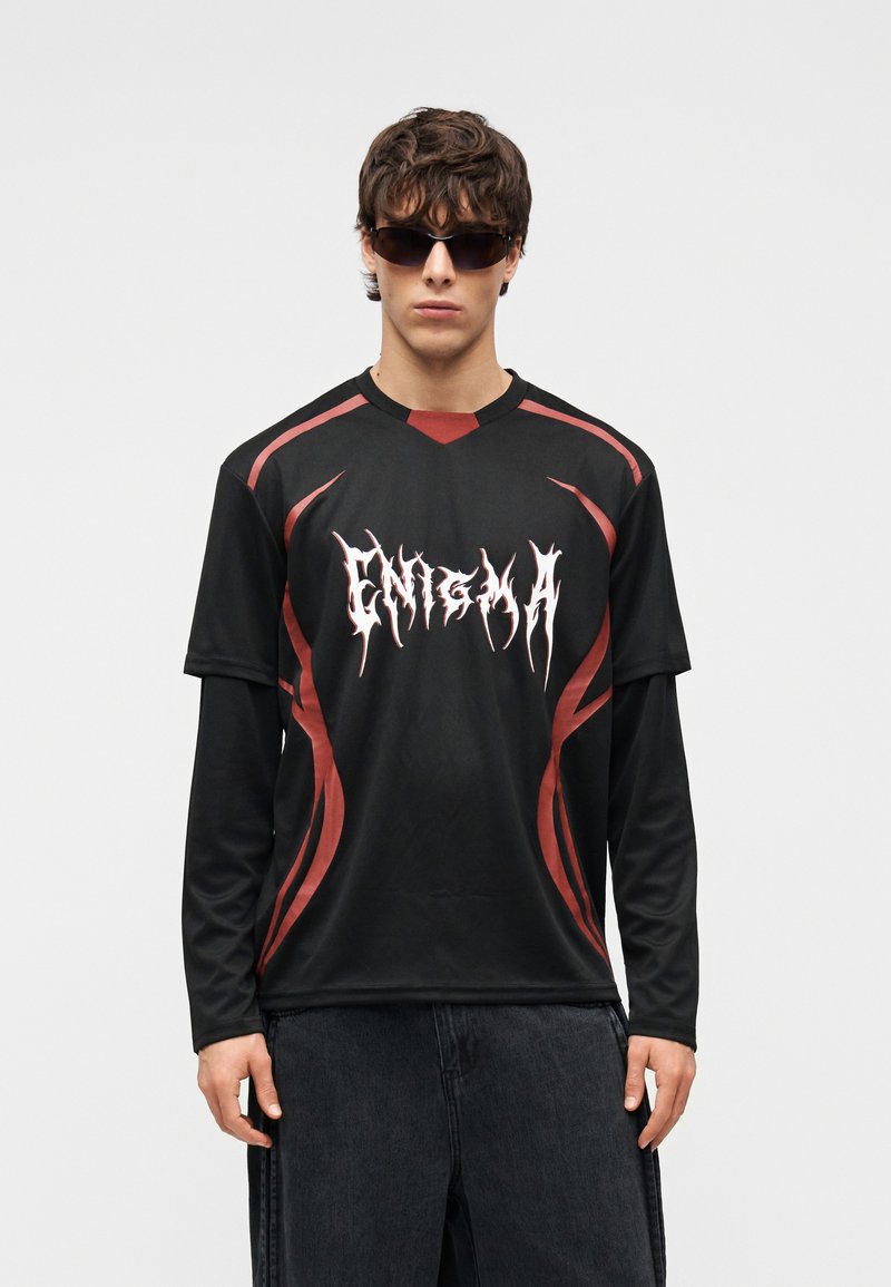 Νεαρός άνδρας που φορά μαύρο μακρυμάνικο πουκάμισο με κόκκινες λεπτομέρειες και λευκή επιγραφή «ENIGMA», σκούρα γυαλιά ηλίου και σκούρο παντελόνι, στέκεται μπροστά σε λευκό φόντο.