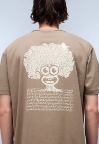 Light brown T-shirt featuring a large white graphic of a tree with a face and a small bird, set against a textured grass-like pattern.