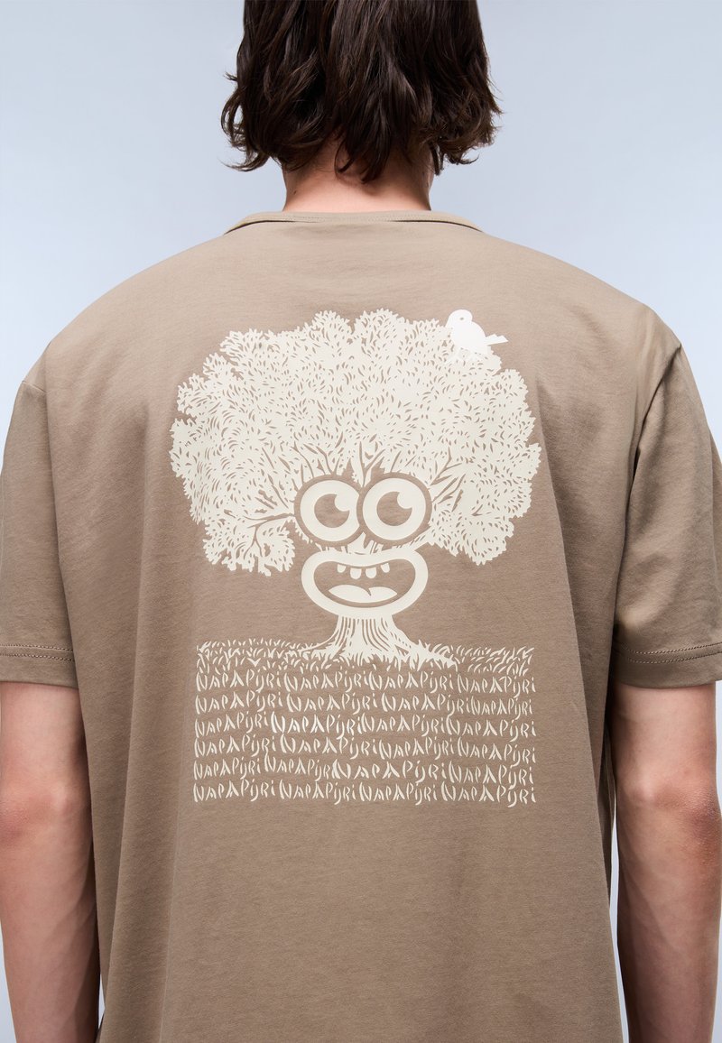 Jasnobrązowy T-shirt z dużą białą grafiką przedstawiającą drzewo z twarzą i małym ptakiem, umieszczoną na tle teksturowanego wzoru przypominającego trawę.