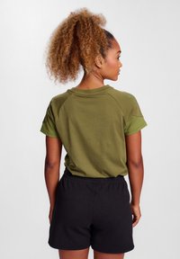 Olivengrøn kortærmet skjorte med en afslappet pasform og sorte shorts. Skjorten har en rund halsudskæring og raglanærmer.
