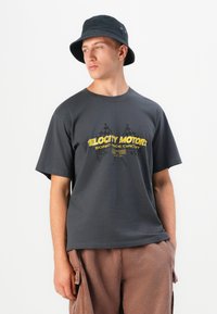 Ung mand iført en sort bucket hat, en mørkegrå grafisk t-shirt med gul tekst og krydsede flag samt brune cargo-bukser mod en hvid baggrund.