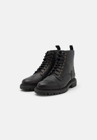 Botas de cuero negro hasta el tobillo con punta redonda, cierre de cordones en la parte delantera, ojales dorados y suela de goma texturizada. Incluye una pestaña en el talón para facilitar su uso.