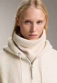 Kremna zip-up kapuca s mehko, puhasto teksturo, ki ima rebrast, visok ovratnik in srebrno zadrgo.