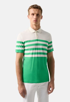 Mężczyzna w białej i zielonej paskowanej koszulce polo oraz białych spodniach, stojący na tle jasnego, jednolitego tła.