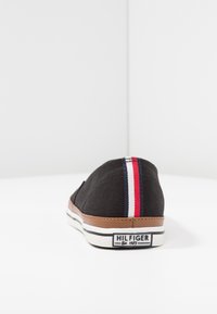Scarpa slip-on nera in tela con rifiniture marroni, suola in gomma bianca e striscia rossa, bianca e blu sul tallone con etichetta "Hilfiger Est 1985".