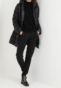 Cappotto piumino nero con cappuccio, finitura testurizzata, abbinato a un maglione nero, pantaloni neri su misura e stivaletti neri alla caviglia con dettagli in oro.