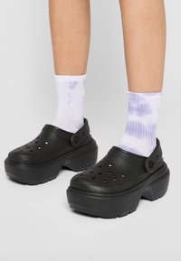 Sabots noirs à plateforme avec des trous de ventilation et une sangle. Portés avec des chaussettes blanches à motif tie-dye violet clair.
