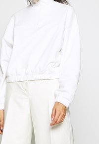 Person med en vit kvartdraget sweatshirt med elastiska ärmslut och nederkant, tillsammans med vita högmidjade byxor framför en neutral bakgrund.