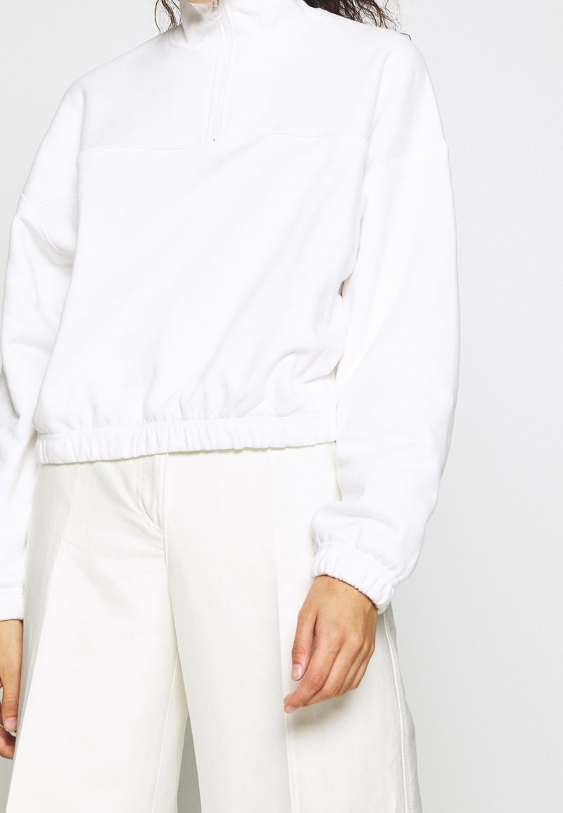 Person med en vit kvartdraget sweatshirt med elastiska ärmslut och nederkant, tillsammans med vita högmidjade byxor framför en neutral bakgrund.