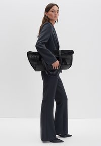 Sac fourre-tout en mesh noir avec un fond solide, doté de deux longues poignées. Le sac a un design structuré et est tenu par une personne en costume sombre.