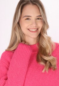 Félfényes rózsaszín kötött pulóver ribbelt textúrával. Hosszú ujjú, laza szabású. A haj hullámos és szőke, oldalt választva.
