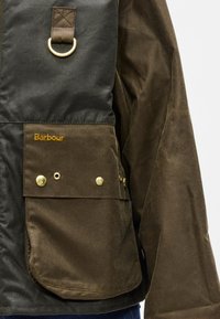 Brown waxed jacket with black accents, gold hardware, visible stitching, and a chest pocket featuring the brand name embroidered in gold.