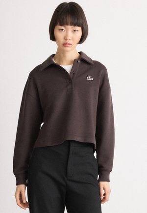 Brun cropped genser laget av mykt materiale med knappestolpe, enkelt brystlogo og lange ermer, kombinert med svarte bukser.
