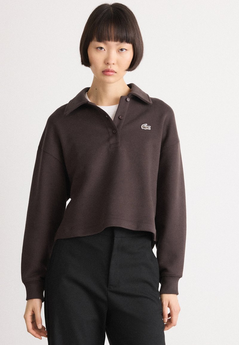 Brown cropped pullover made of soft material with a buttoned collar, single chest logo, and long sleeves, paired with black pants.