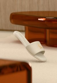 Λευκό σανδάλι slide από λείο υλικό, με φαρδιά λουράκι και χρυσή διακοσμητική λεπτομέρεια. Τοποθετημένο σε απαλό, ουδέτερο φόντο.