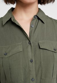 Krátký rukáv košile v olivově zelené látce, s knoflíkovou légu, dvěma náprsními kapsami a kontrastními navy knoflíky. Hladká textura.