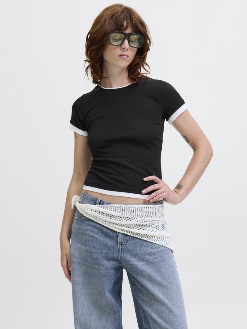 Fiatal személy hullámos barna hajjal, túlságosan nagy napszemüveget visel, fekete, fehér szegélyű szűkített pólót, világoskék farmert és fehér hálós pulóvert, amelyet a derékra kötött.