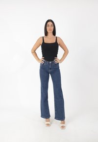 Czarny dopasowany top na ramiączkach, niebieskie szerokie dżinsy z wysokim stanem, fakturowany materiał oraz białe sandały na obcasie, stojąca na białym tle.