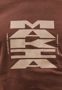 Pruun T-särk, millel on suur beež graafiline tekst "MAYAH" ja "HELSINKI EST. 2003." Pehme puuvillane materjal ja lõdva lõikega.