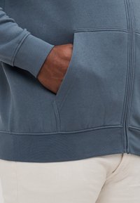 Blue cotton hoodie with a zip front, featuring side pockets and ribbed cuffs. The material has a smooth texture, with visible stitching details.