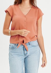 Blouse couleur rouille à manches courtes, col en V et boutonnée devant. Dotée d'un lien à la taille et d'un tissu texturé avec un motif subtil.
